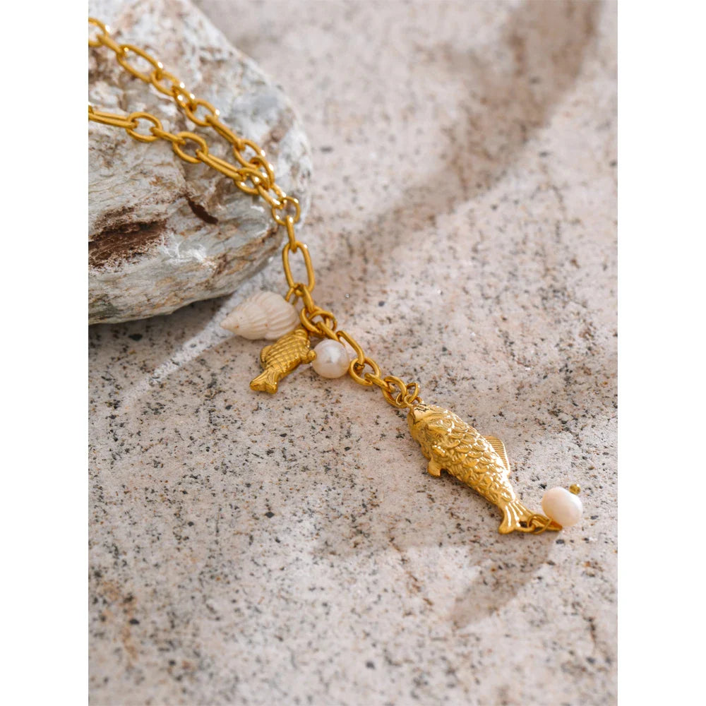 Gold-tone coastal necklace with fish pendant, shell charm, and pearl charms on sandy surface.